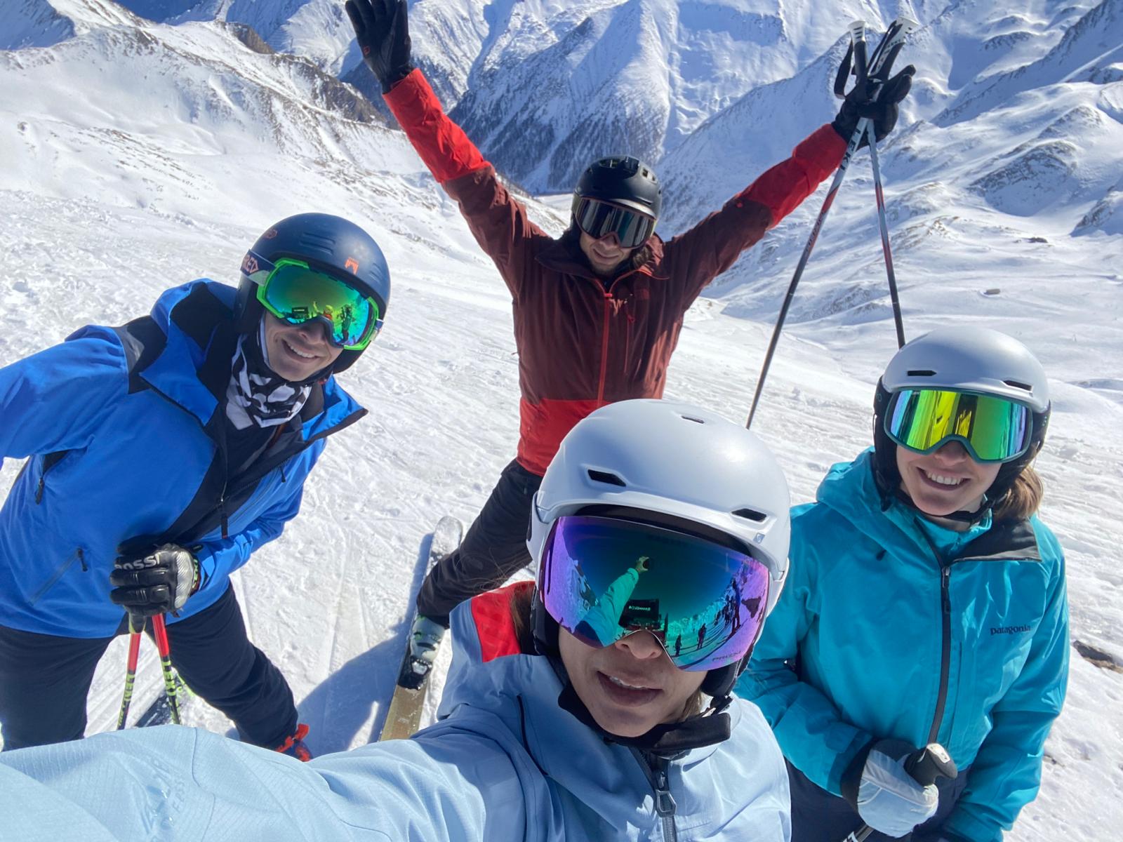 Four adult skiers taking a group selfie on a piste with snow-covered mountains behind them