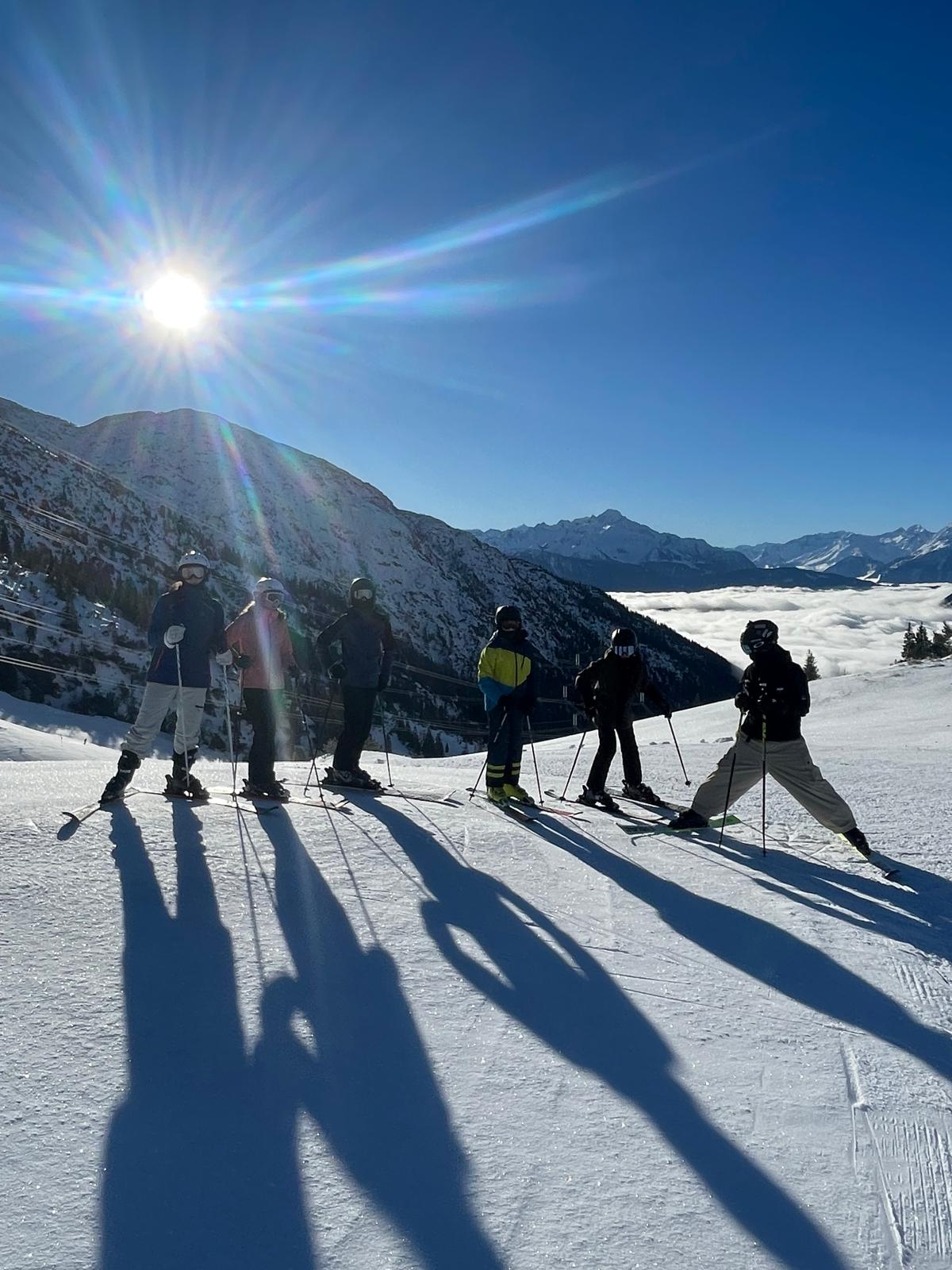 Six skiers standing in a line on a ridge, silhouetted against a low sun above a sea of cloud
