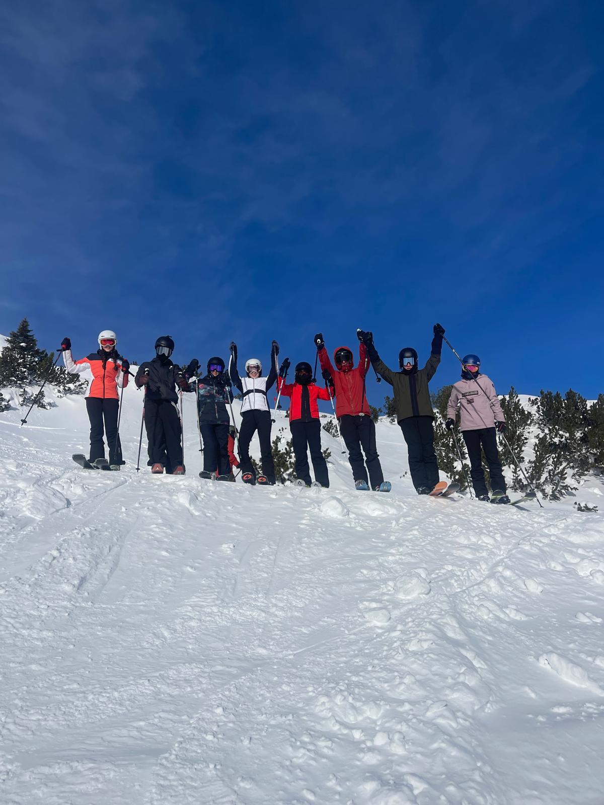 Eight teenage skiers and snowboarders standing in a line with poles raised under a deep blue sky