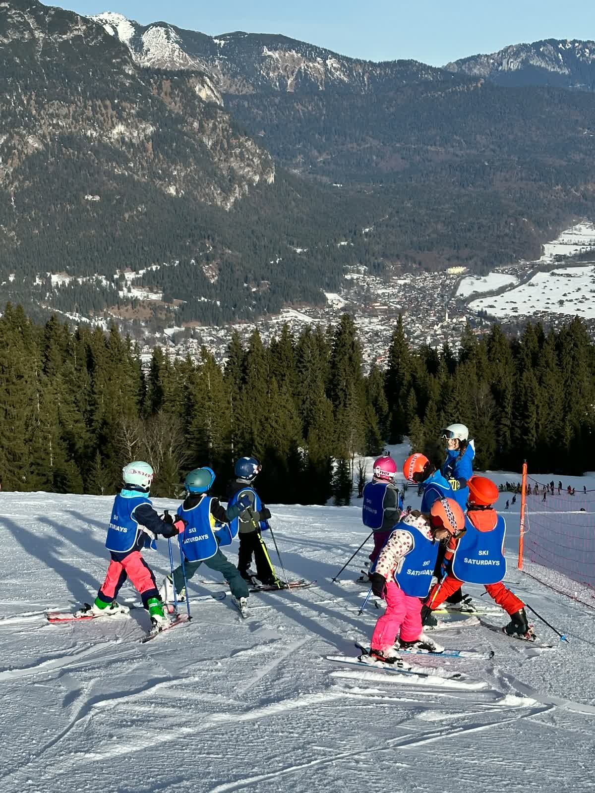 Young children in Ski Saturdays bibs gathered with their instructor at the top of a piste, with a snowy valley town and forested mountains below