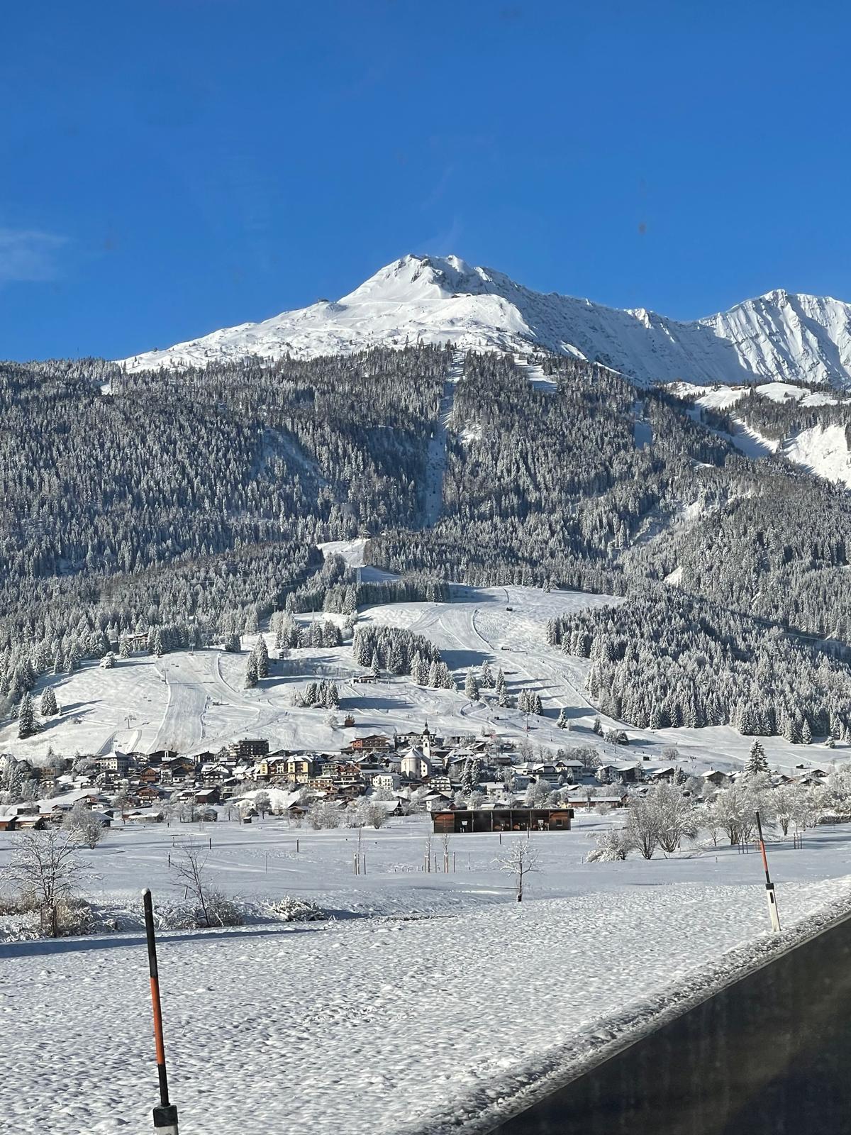 An alpine village below wooded slopes and a snow-covered peak on a clear winter morning